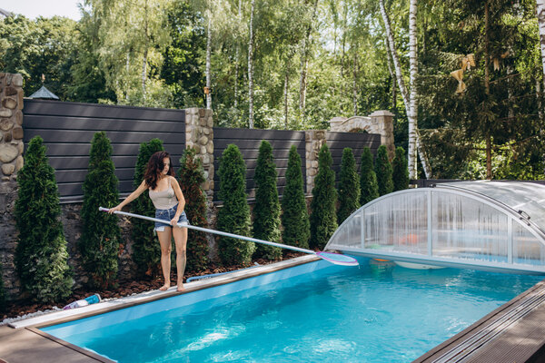 Eine Frau steht am Pool und entfernt mit einem Netz Blätter aus dem Wasser, während sie den Pool mit Abdeckung reinigt.